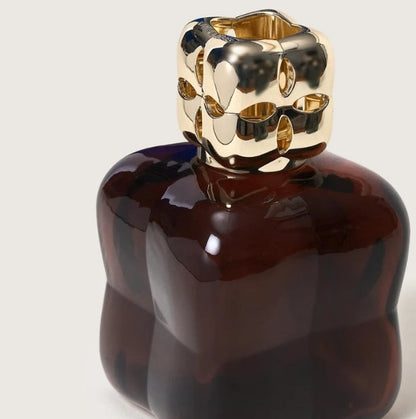 A dark prune glossy glass bottle with a sculptural, wavy form and a highly reflective gold metallic cap.