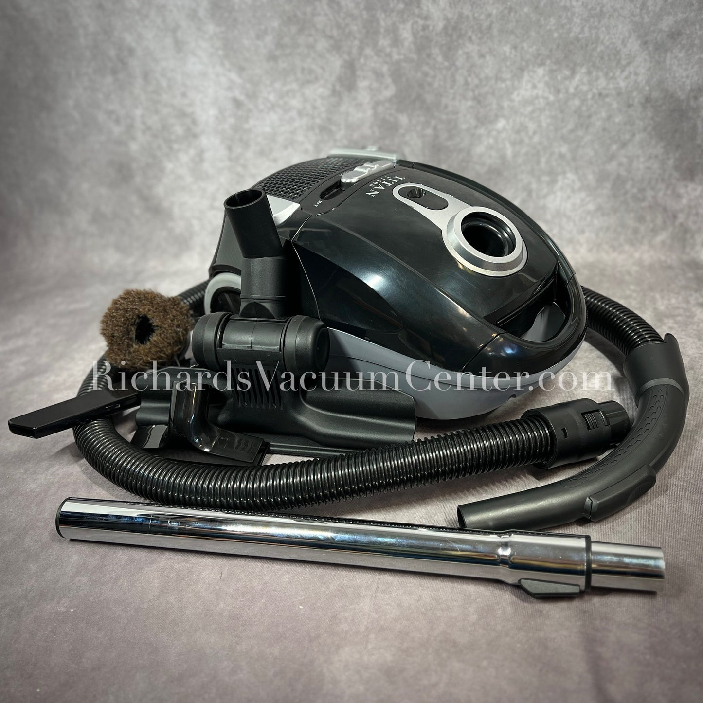 A black Titan T1400 deluxe canister vacuum with a telescopic wand, power cord, and premium accessory tools displayed.