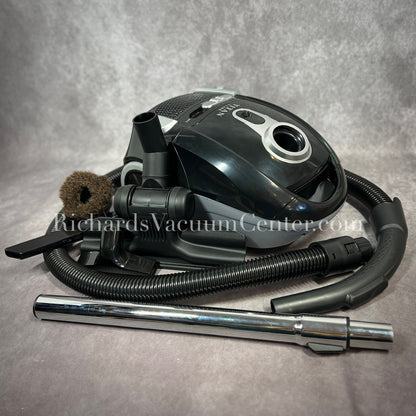 A black Titan T1400 deluxe canister vacuum with a telescopic wand, power cord, and premium accessory tools displayed.